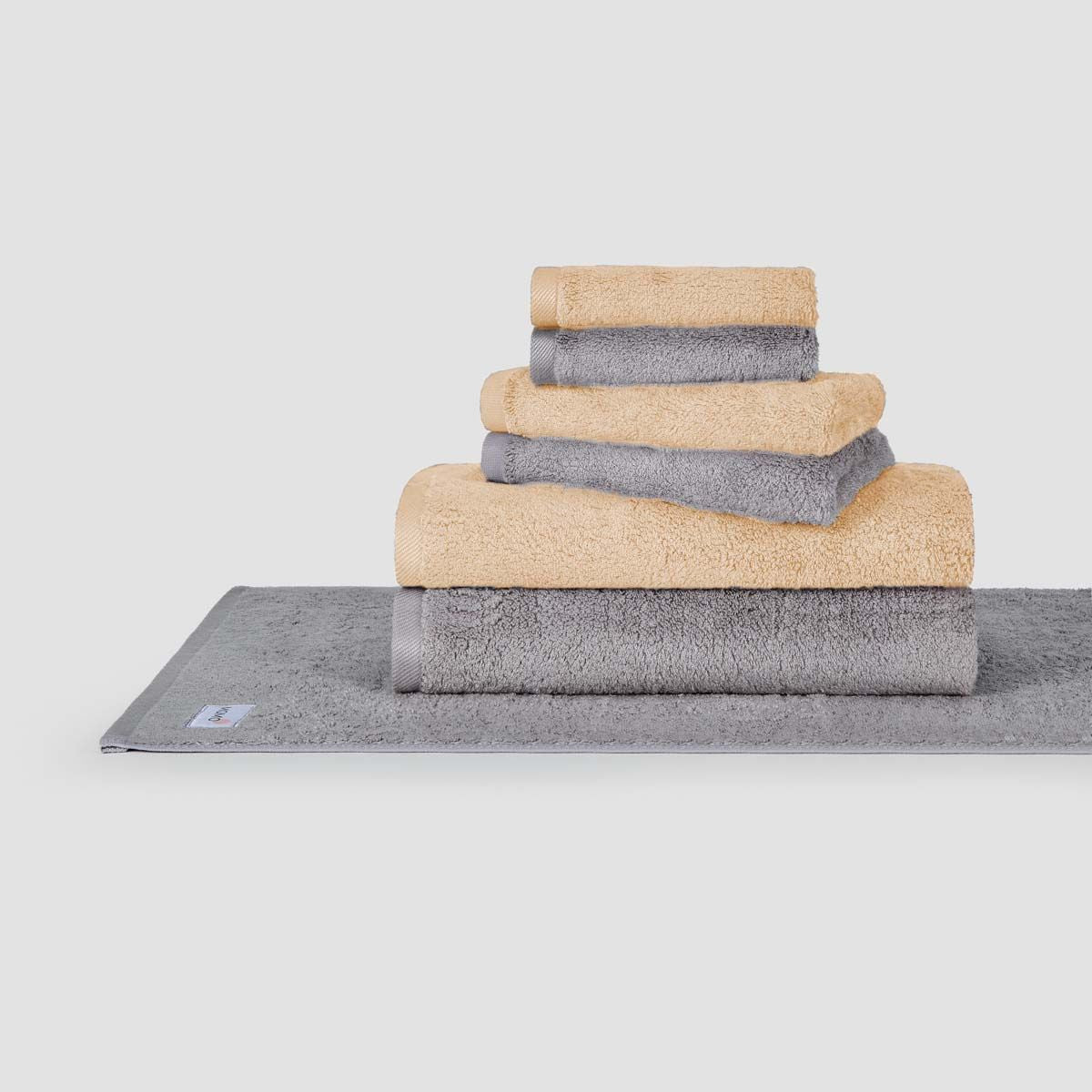 Set di asciugamani impilati beige e grigio Momo Cotton esposto su tappetino da bagno grigio su sfondo bianco