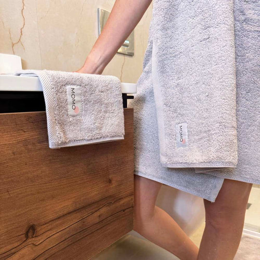 Set di asciugamani in cotone Momo esposto su scaffale in legno da bagno con persona che tiene asciugamani beige piegati