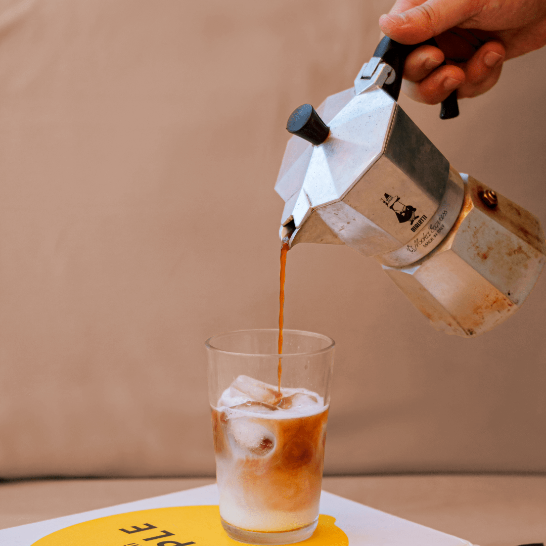 Hot moka being poured into a glass cup with ice on a beige background.