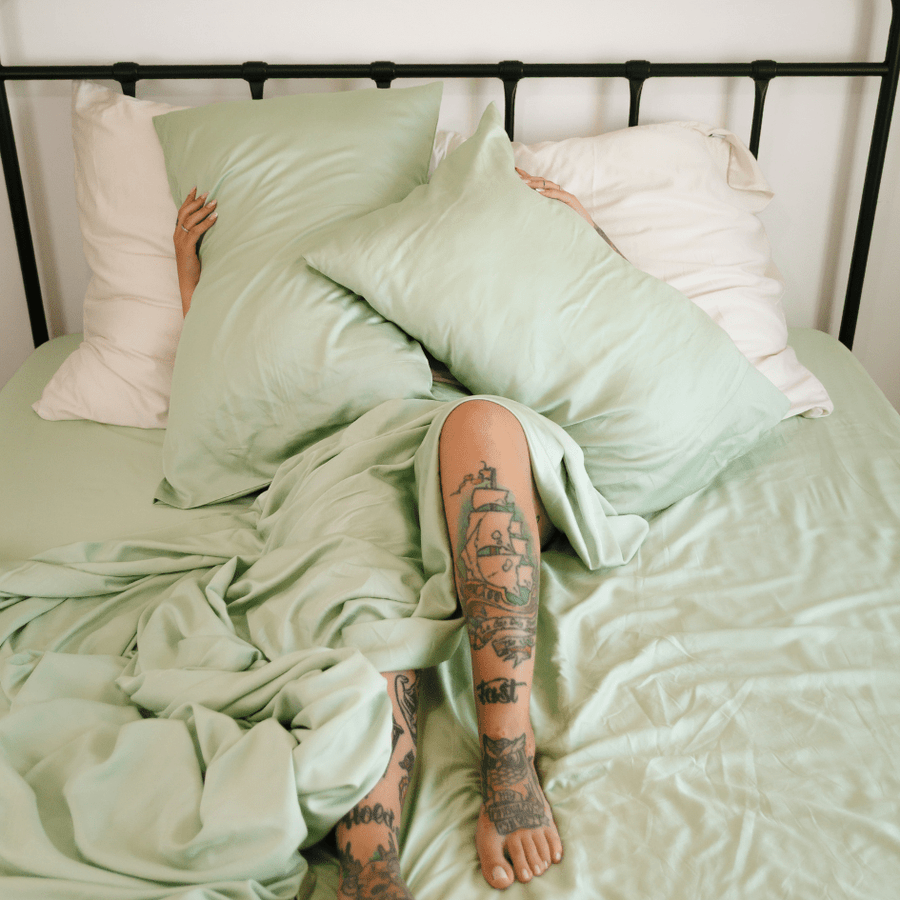 Persona sdraiata nel letto che indossa biancheria da letto in bambù verde e bianco con tatuaggi visibili sulle gambe