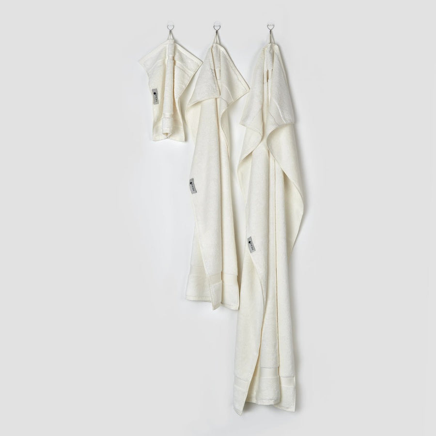 Tre asciugamani in bambù crema appesi a ganci che mostrano le taglie viso, mano e bagno della collezione Alba di momo-cotton