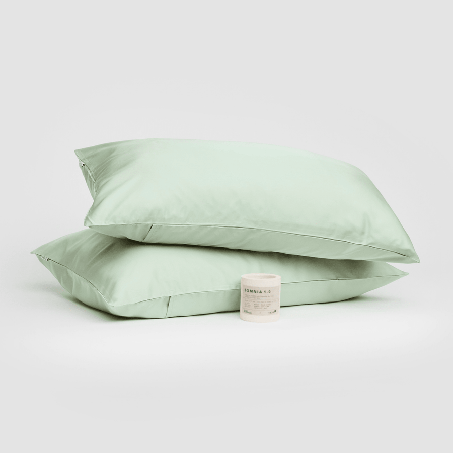 Coppia di federe in cotone verde salvia impilate con candela Somnia 1.0 su sfondo bianco