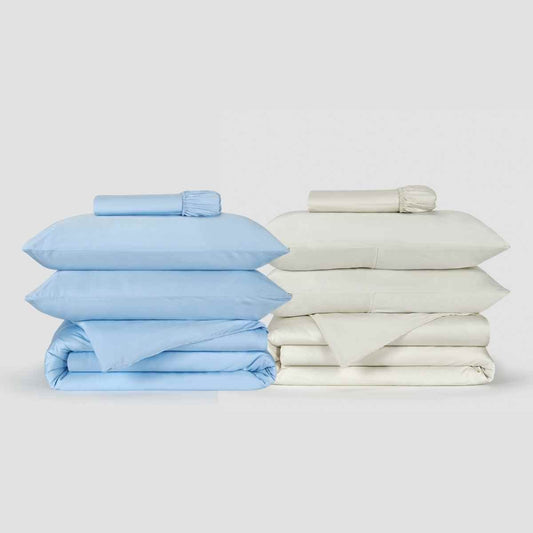 Due completi di copripiumino Momo Cotton affiancati, uno blu chiaro e uno crema, con lenzuola piegate e biancheria arrotolata